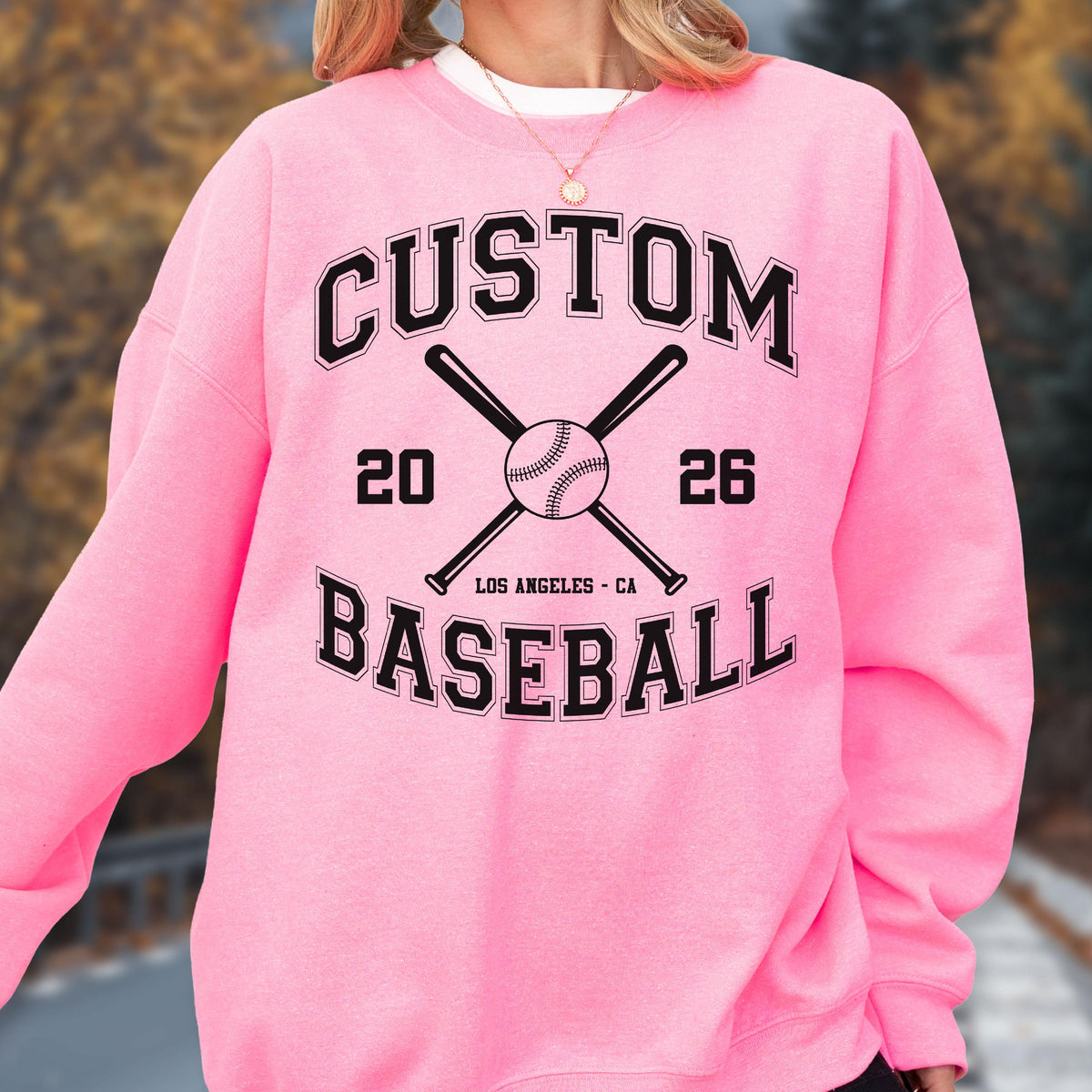 May include: A bright pink crewneck sweatshirt featuring a black graphic print. The design includes the text CUSTOM BASEBALL, the numbers 20 and 26, two crossed baseball bats, a baseball, and the text LOS ANGELES - CA in a collegiate font style.