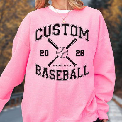 May include: A bright pink crewneck sweatshirt featuring a black graphic print. The design includes the text CUSTOM BASEBALL, the numbers 20 and 26, two crossed baseball bats, a baseball, and the text LOS ANGELES - CA in a collegiate font style.