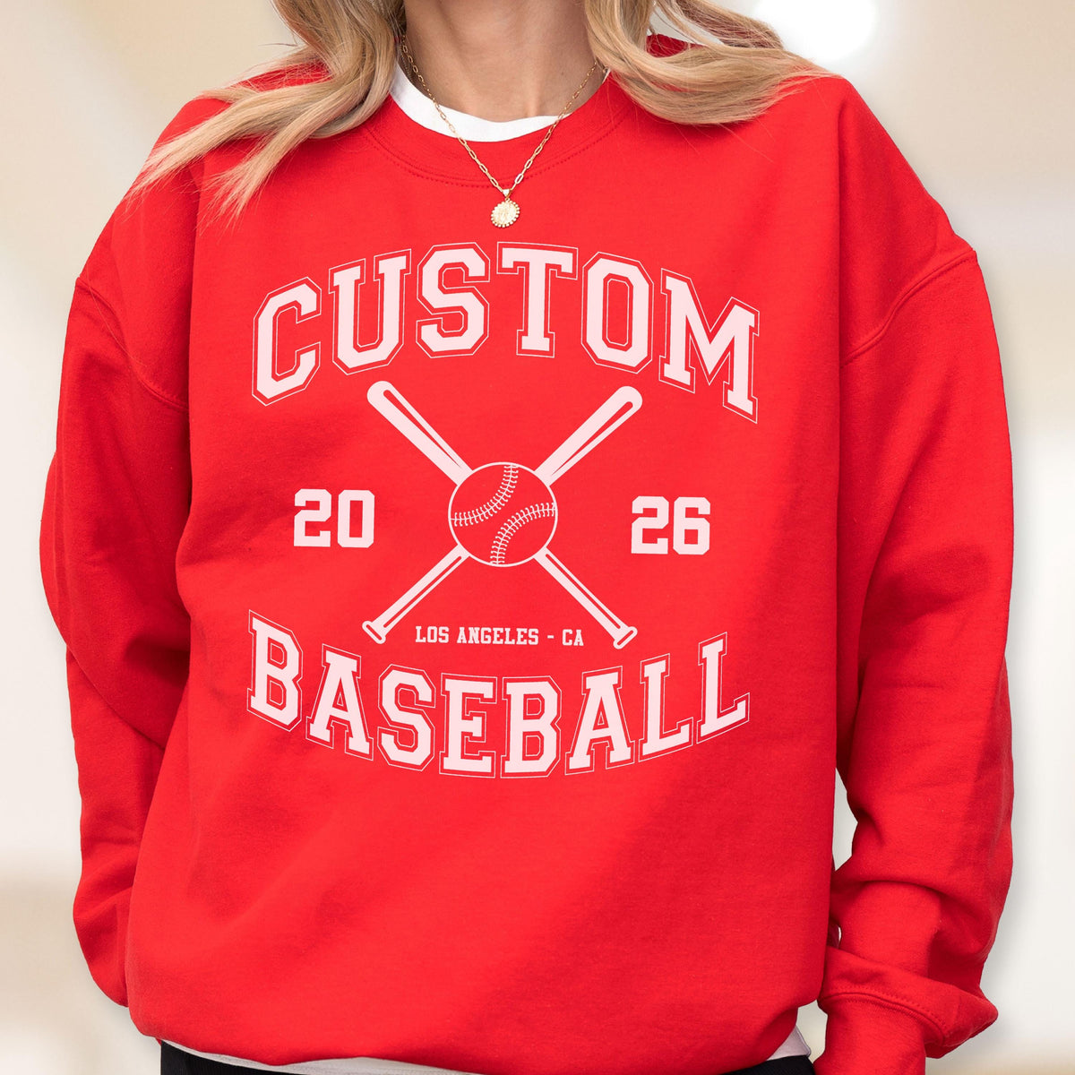 May include: A bright red crewneck sweatshirt featuring a white graphic print. The design includes the text CUSTOM BASEBALL, the numbers 20 and 26, two crossed baseball bats, a baseball, and the text LOS ANGELES - CA in a collegiate font style.