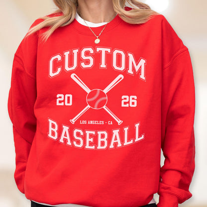May include: A bright red crewneck sweatshirt featuring a white graphic print. The design includes the text CUSTOM BASEBALL, the numbers 20 and 26, two crossed baseball bats, a baseball, and the text LOS ANGELES - CA in a collegiate font style.
