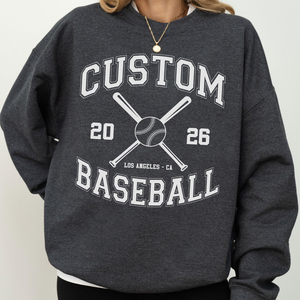 May include: A dark charcoal gray crewneck sweatshirt featuring white screen-printed text that reads CUSTOM BASEBALL, the numbers 20 and 26, and the location LOS ANGELES - CA. The design includes a graphic of two crossed baseball bats and a ball.