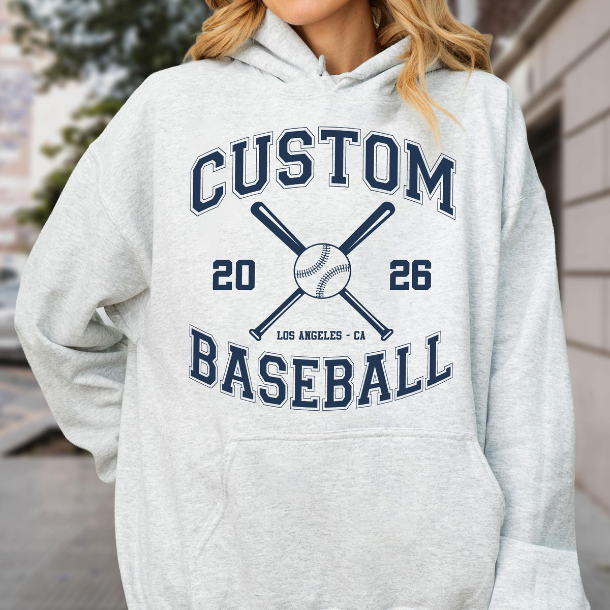 May include: A light grey hooded sweatshirt featuring a dark blue graphic print. The design includes the text CUSTOM BASEBALL, the numbers 20 and 26, the location LOS ANGELES - CA, and an illustration of a baseball centered between two crossed bats.