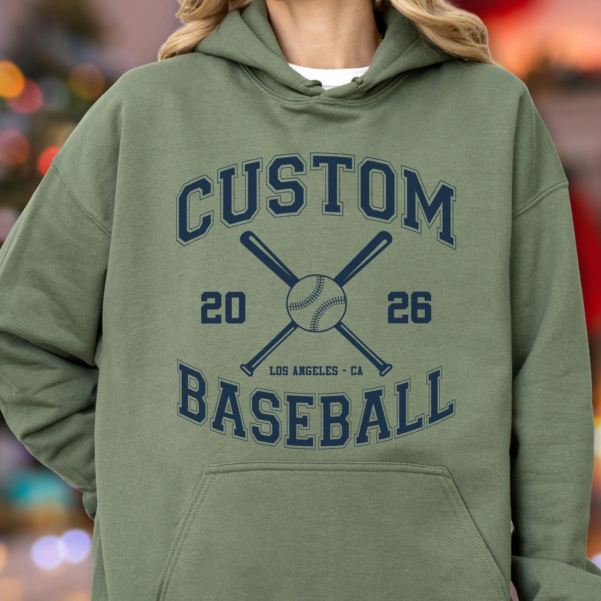 May include: An olive green hooded sweatshirt featuring a dark navy blue graphic print. The design includes the text CUSTOM BASEBALL, the numbers 20 and 26, two crossed baseball bats, a baseball, and the text LOS ANGELES - CA in the center.