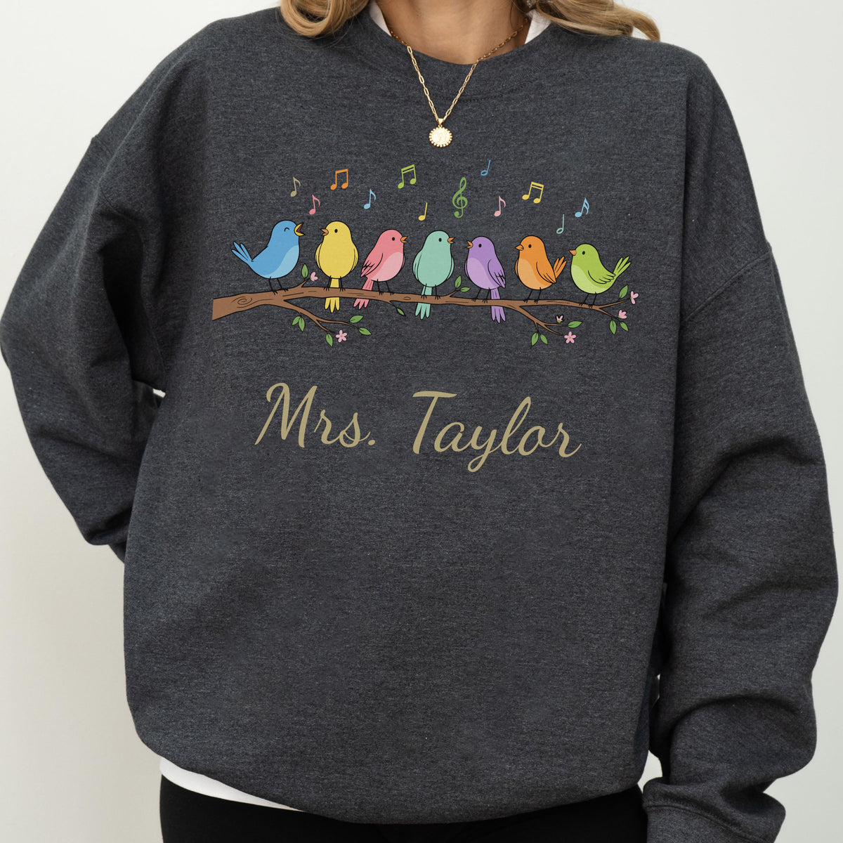 May include: A dark charcoal gray crewneck sweatshirt featuring a graphic of seven colorful birds perched on a brown branch with musical notes above them. The text "Mrs. Taylor" is printed in a gold script font below the bird illustration.