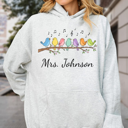 May include: A light gray hooded sweatshirt featuring a graphic of seven colorful birds perched on a brown branch with musical notes above them. The text Mrs. Johnson is printed in black script below the branch illustration on the front.