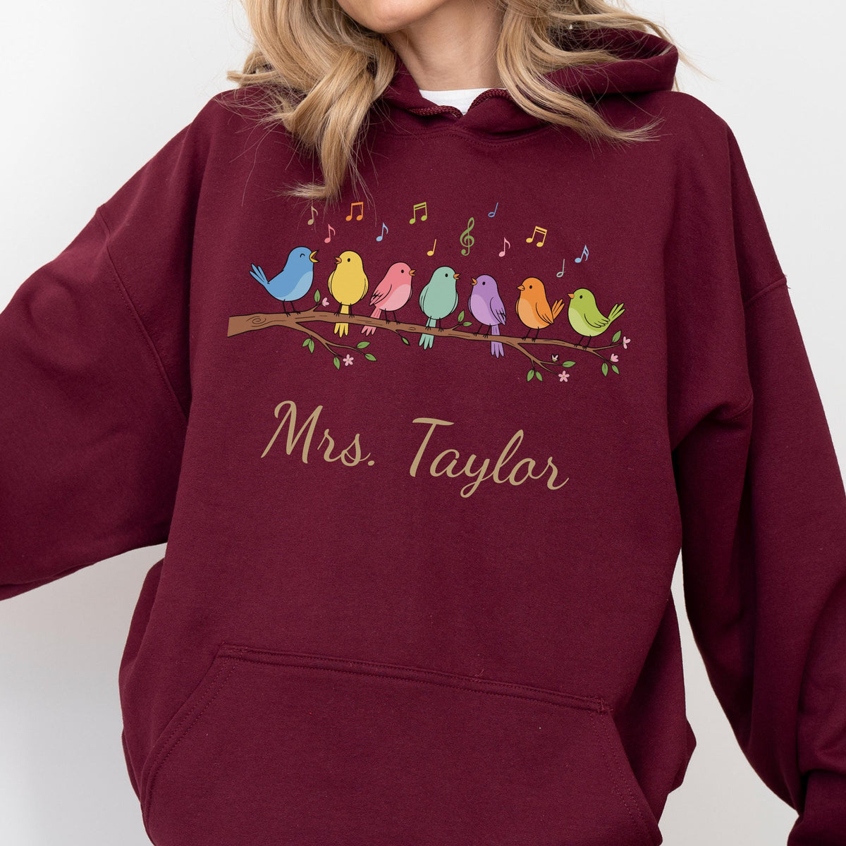 May include: A maroon hooded sweatshirt featuring a graphic of seven colorful birds perched on a brown branch with musical notes above them. Below the birds, the text "Mrs. Taylor" is printed in a cursive, light-colored script font.