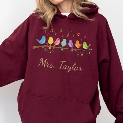 May include: A maroon hooded sweatshirt featuring a graphic of seven colorful birds perched on a brown branch with musical notes above them. Below the birds, the text "Mrs. Taylor" is printed in a cursive, light-colored script font.