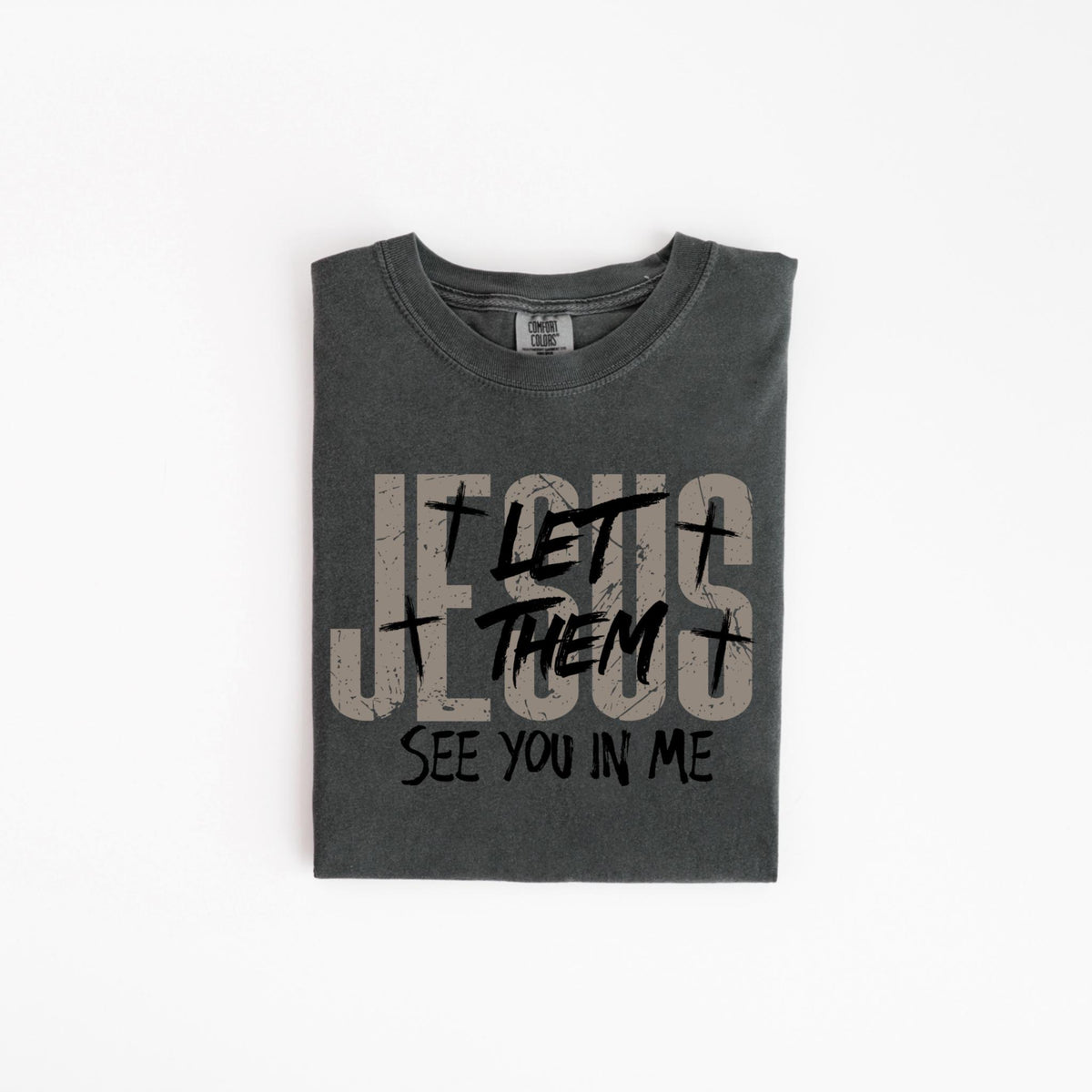 May include: Dark gray t-shirt with the text "JESUS LET THEM SEE YOU IN ME" in a distressed font. The shirt has a crew neck and is folded neatly. The text includes cross symbols.