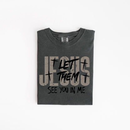 May include: Dark gray t-shirt with the text "JESUS LET THEM SEE YOU IN ME" in a distressed font. The shirt has a crew neck and is folded neatly. The text includes cross symbols.