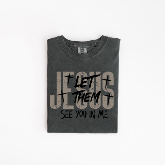 May include: Dark gray t-shirt with the text "JESUS LET THEM SEE YOU IN ME" in a distressed font. The shirt has a crew neck and is folded neatly. The text includes cross symbols.