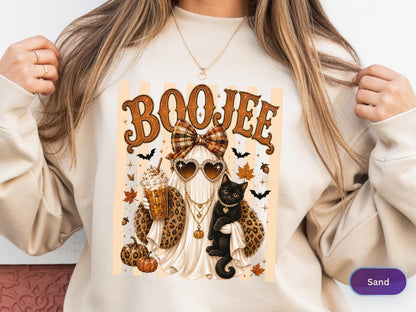 May include: A sand-colored sweatshirt featuring a graphic print of a ghost wearing heart-shaped sunglasses, a plaid bow, and a leopard print shawl. The ghost holds an iced coffee and a black cat, surrounded by pumpkins, autumn leaves, and bats.
