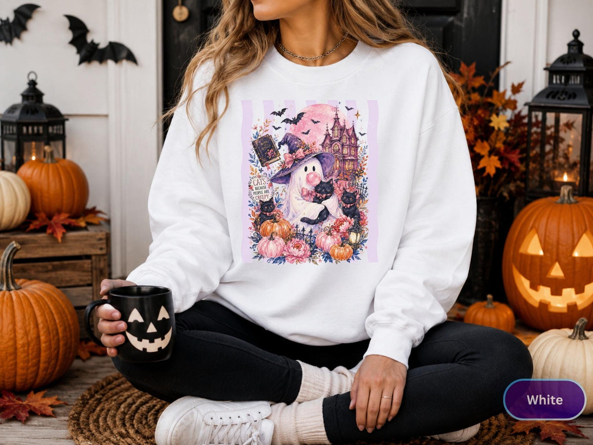 May include: A white long-sleeve sweatshirt featuring a graphic print of a ghost wearing a witch hat, holding black cats, surrounded by pumpkins, flowers, a haunted house, and bats. The text on the graphic reads Cats, Books, and Creepy.