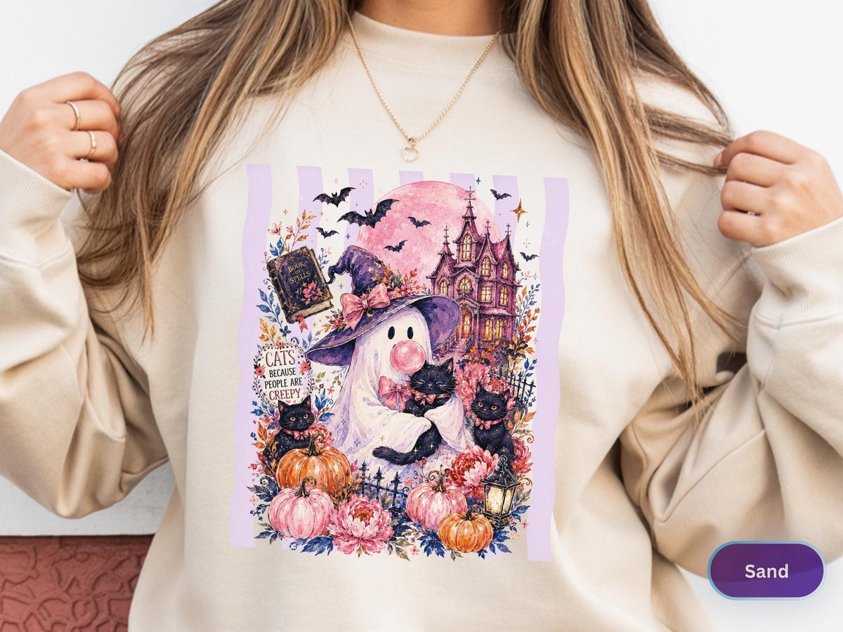 May include: A sand-colored sweatshirt featuring a graphic print of a ghost wearing a purple witch hat and blowing a pink bubble. The design includes black cats, pumpkins, a haunted house, bats, and the text "Cats because people are creepy."