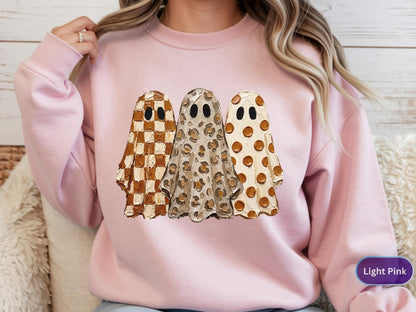 May include: A light pink crewneck sweatshirt featuring a graphic of three ghosts. The left ghost has a brown and white checkered pattern, the middle ghost has a brown leopard print, and the right ghost has a brown polka dot pattern.