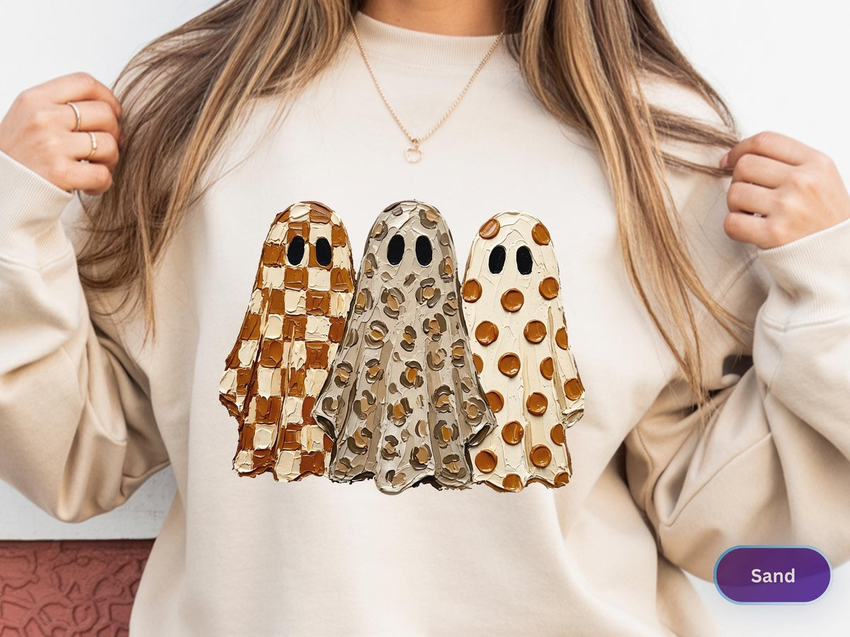 May include: A sand-colored crewneck sweatshirt featuring a graphic of three ghosts with textured, paint-like brushstrokes. The left ghost has a brown and white checkerboard pattern, the center has a leopard print, and the right has brown polka dots.