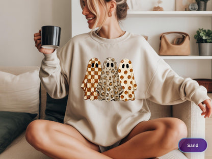 May include: A sand-colored crewneck sweatshirt featuring a graphic print of three ghosts standing side-by-side. The first ghost has a brown and white checkered pattern, the second has a leopard print, and the third has a brown polka dot pattern.