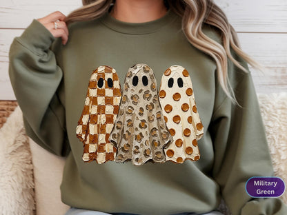 May include: A military green crewneck sweatshirt featuring a graphic of three ghosts with black oval eyes. The left ghost has a brown and cream checkerboard pattern, the middle ghost has a brown and cream leopard print, and the right ghost has brown polka dots.
