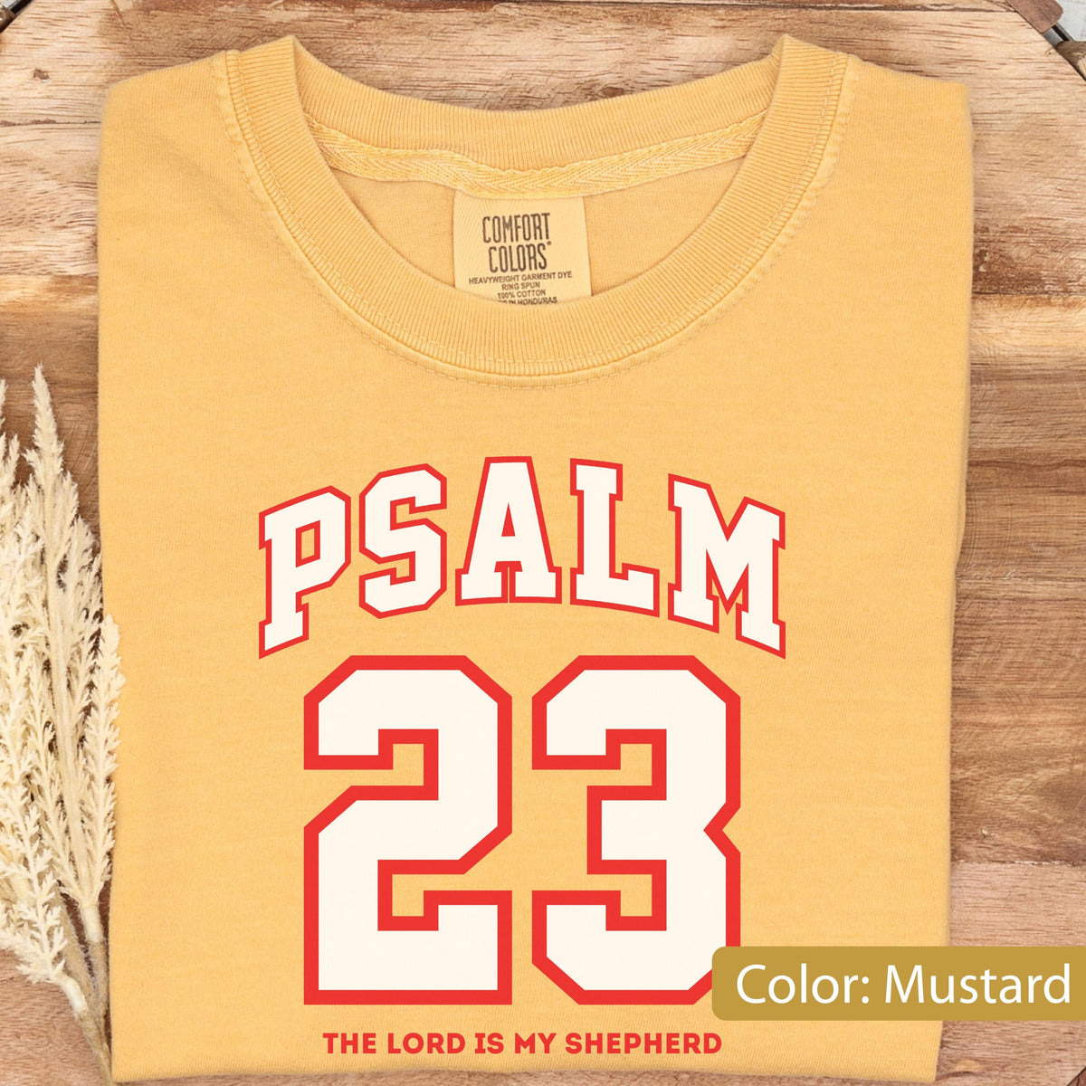 May include: A mustard yellow crew neck t-shirt featuring the text PSALM 23 in a white collegiate font with a red outline. Below the number 23, the text reads THE LORD IS MY SHEPHERD in red. The shirt has a Comfort Colors brand label at the neck.