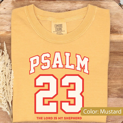 May include: A mustard yellow crew neck t-shirt featuring the text PSALM 23 in a white collegiate font with a red outline. Below the number 23, the text reads THE LORD IS MY SHEPHERD in red. The shirt has a Comfort Colors brand label at the neck.