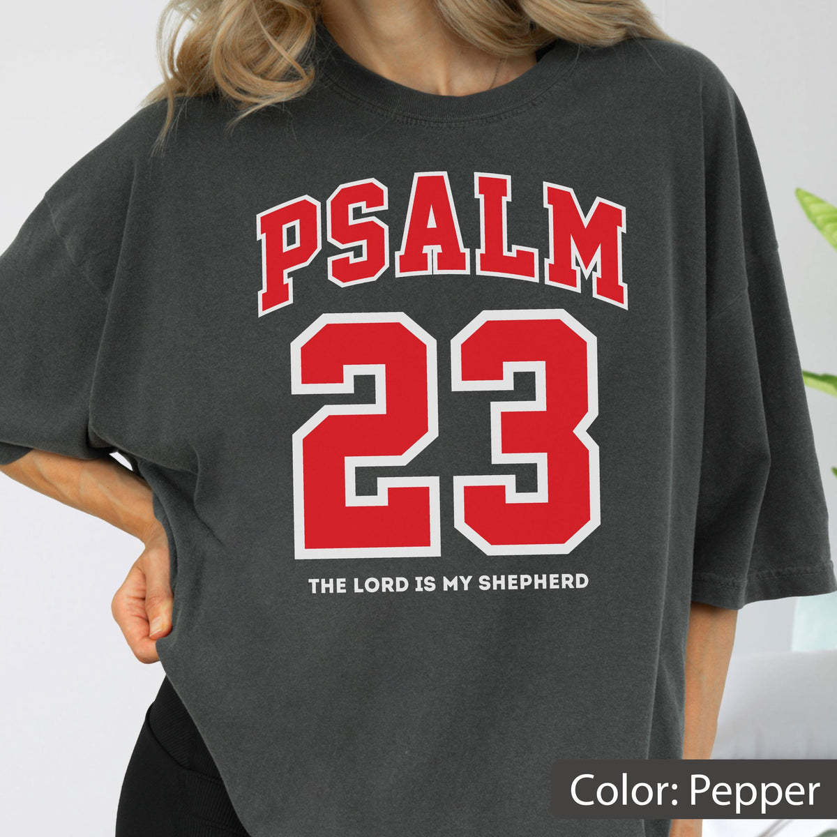 May include: A charcoal gray short-sleeve t-shirt featuring the red text PSALM 23 in a collegiate font style, with the number 23 in large red block lettering below it and the white text THE LORD IS MY SHEPHERD printed at the bottom.