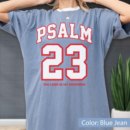 May include: A blue jean colored short-sleeve t-shirt featuring the text PSALM 23 in large white block letters with a red outline. Below the number 23, the text THE LORD IS MY SHEPHERD is printed in red capital letters on the front of the garment.