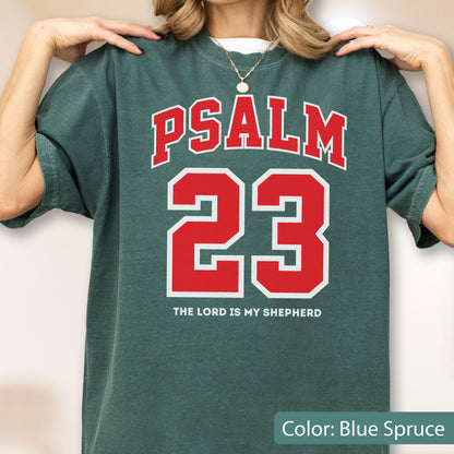 May include: A blue spruce short-sleeve t-shirt featuring a red and white graphic print. The text reads PSALM 23 in a large collegiate font, with the phrase THE LORD IS MY SHEPHERD printed in smaller white capital letters underneath the number 23.