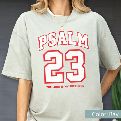 May include: A light sage green short-sleeve t-shirt featuring the red text PSALM 23 in a collegiate font, with the number 23 in large red block lettering below it, and the text THE LORD IS MY SHEPHERD printed in smaller red letters at the bottom.