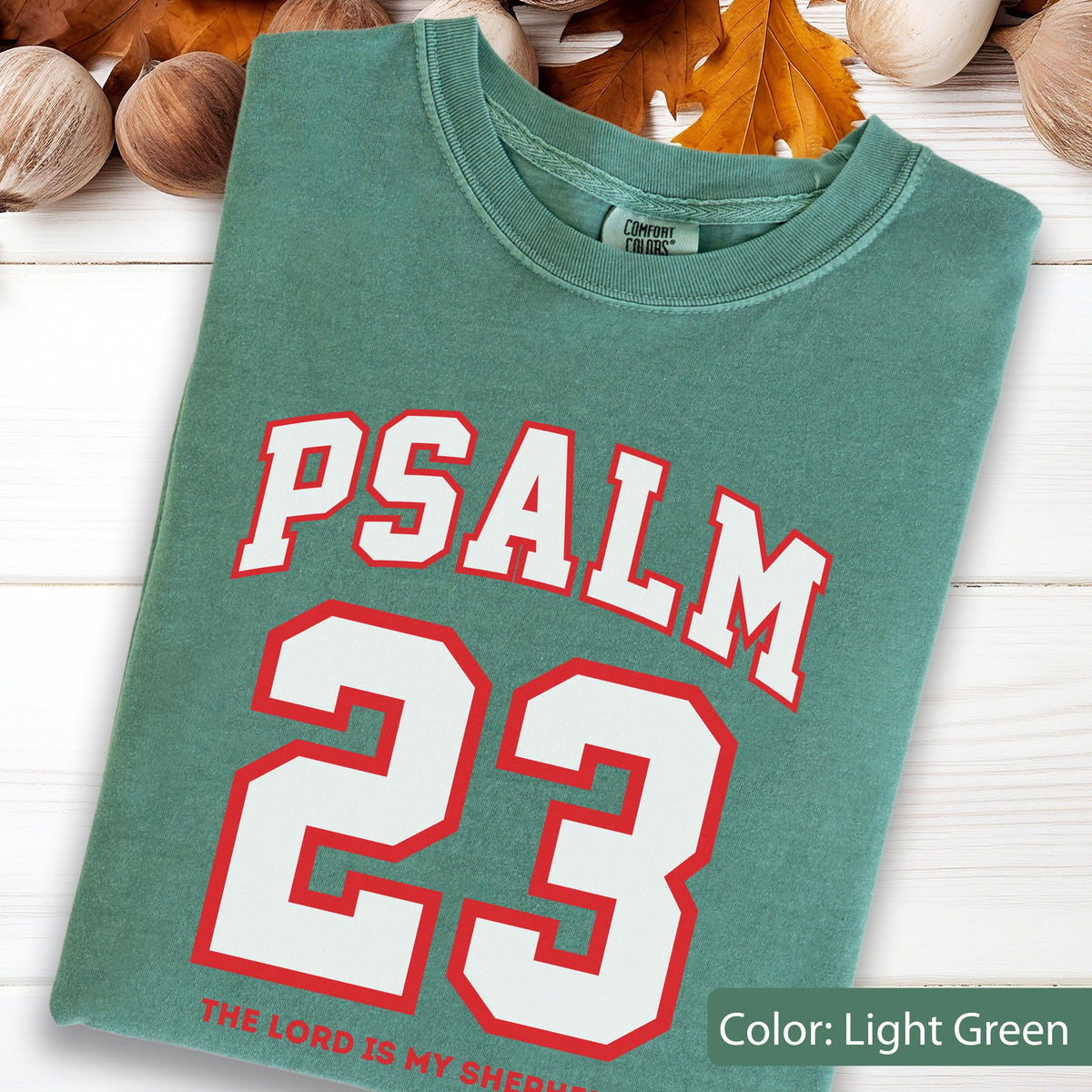 May include: A light green crew neck t-shirt featuring the text PSALM 23 in large white letters with red outlines. Below the number 23, the text THE LORD IS MY SHEPHERD is printed in red. The shirt has a Comfort Colors brand label at the neck.