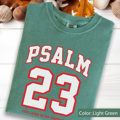 May include: A light green crew neck t-shirt featuring the text PSALM 23 in large white letters with red outlines. Below the number 23, the text THE LORD IS MY SHEPHERD is printed in red. The shirt has a Comfort Colors brand label at the neck.