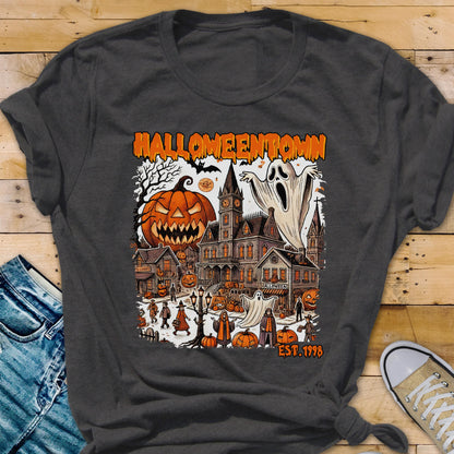 May include: Dark gray t-shirt featuring a Halloween-themed graphic. The design includes a jack-o'-lantern, haunted houses, ghosts, and the text "Halloweentown EST. 1998" in orange. The shirt is made of a soft material.