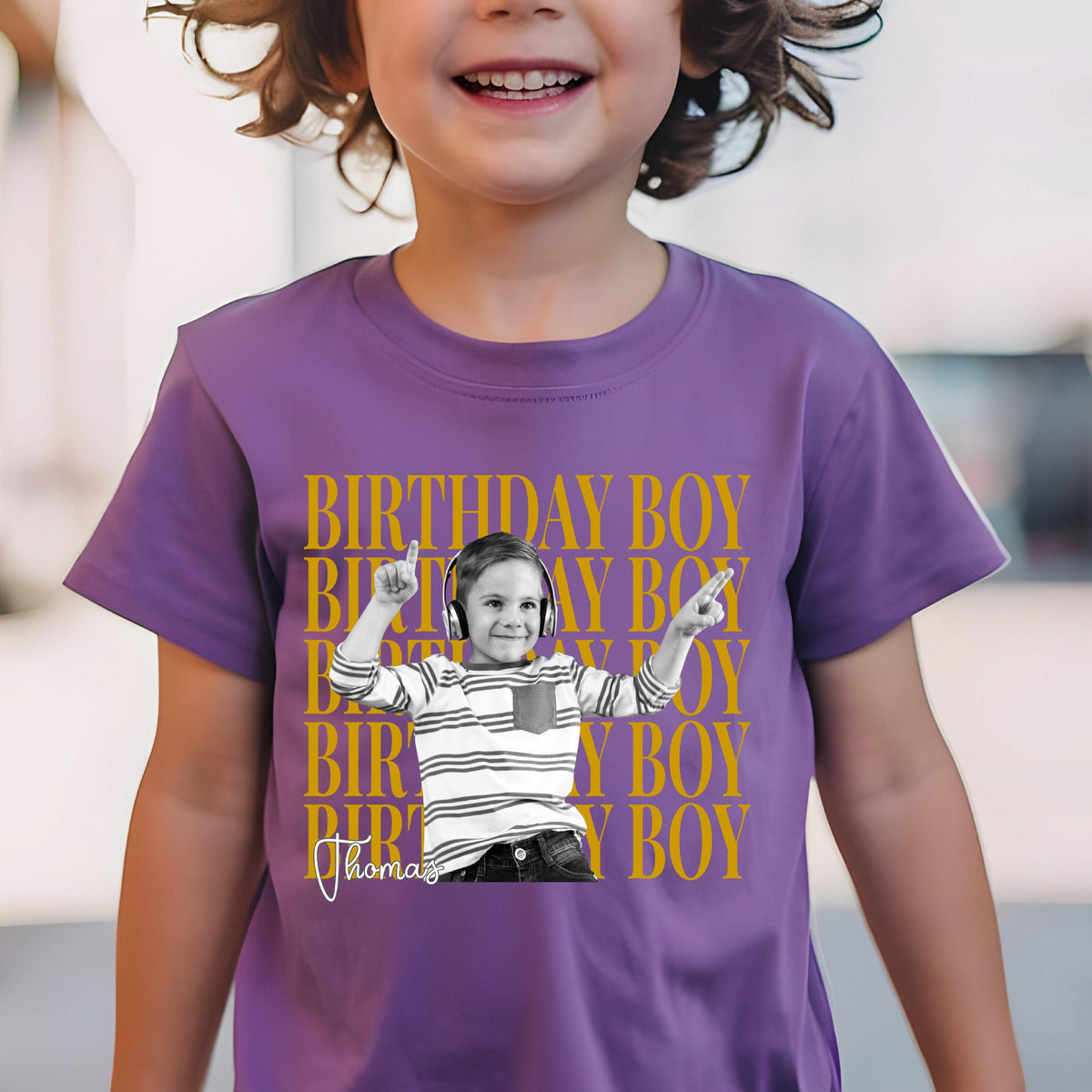May include: A purple short-sleeved t-shirt featuring a repeating gold text pattern that reads BIRTHDAY BOY. Centered is a black and white photograph of a child wearing headphones and a striped shirt, with the name Thomas written in white script below.