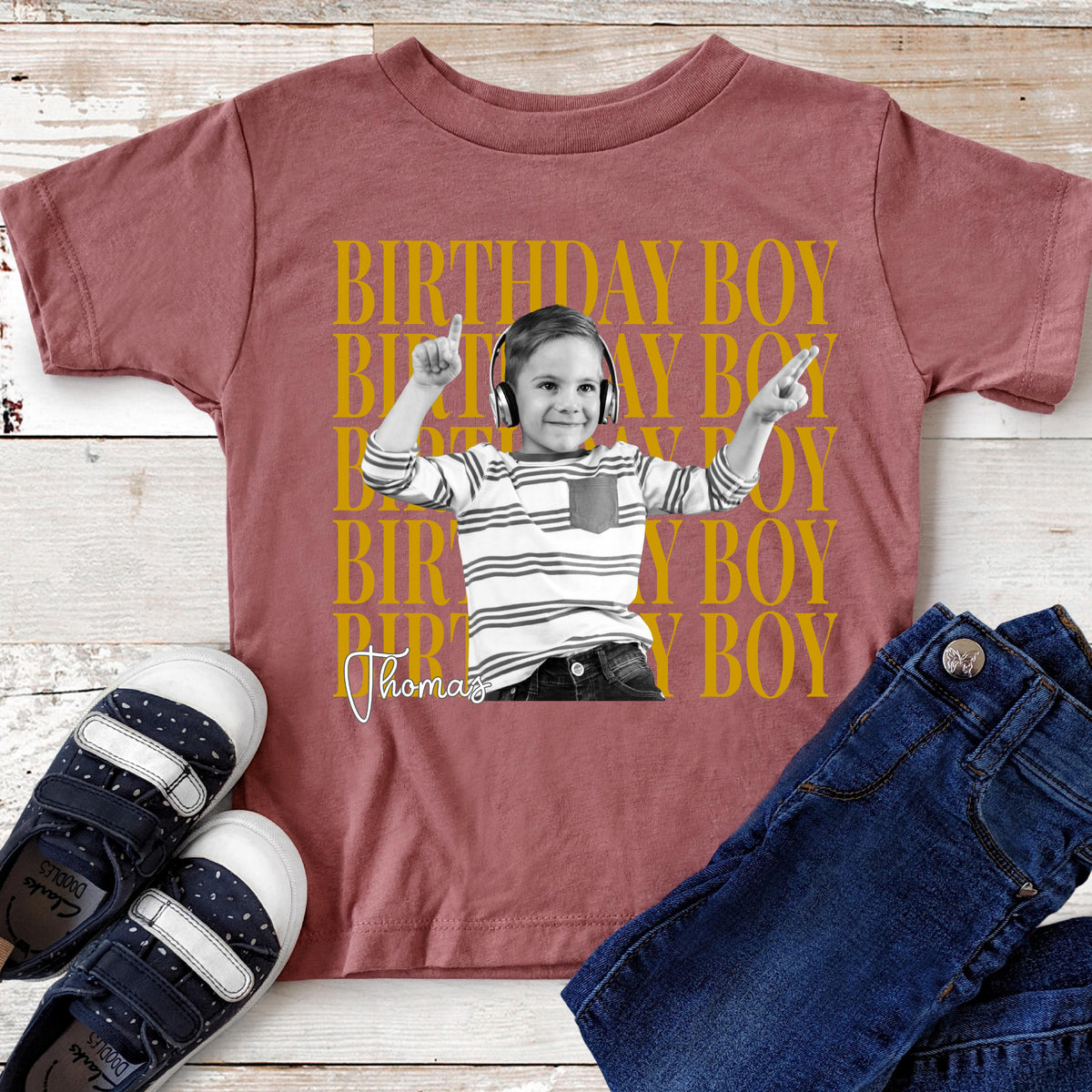 May include: A dusty rose t-shirt featuring a black and white photograph of a child wearing headphones, layered over repeating yellow text that reads BIRTHDAY BOY. The name Thomas is written in white cursive script below the child's image.