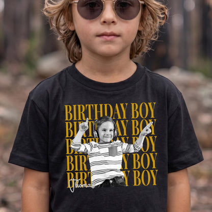 May include: A black short-sleeved t-shirt featuring a repeating yellow text pattern that reads BIRTHDAY BOY. Centered on the shirt is a black and white photograph of a child wearing headphones and a striped shirt, with the name Thomas written below.