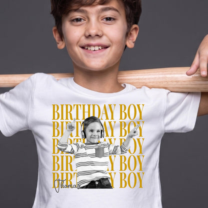 May include: A white t-shirt featuring a repeating gold text pattern that reads BIRTHDAY BOY. Centered is a black and white photograph of a child wearing headphones and a striped shirt, with the name Thomas written in black script below the photo.
