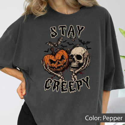 May include: A pepper-colored t-shirt featuring a graphic print of a jack-o'-lantern and a human skull held by skeletal hands. Above and below the central illustration, the text reads STAY CREEPY in a dripping font, surrounded by small bats.