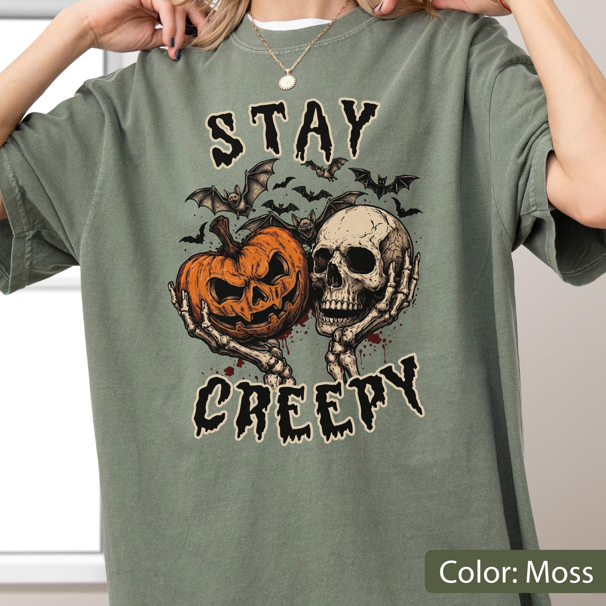 May include: A moss green short-sleeve t-shirt featuring a graphic print of a jack-o'-lantern and a human skull held by skeletal hands. Above and below the illustration, the text reads STAY and CREEPY in a dark, jagged, stylized font.