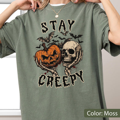 May include: A moss green short-sleeve t-shirt featuring a graphic print of a jack-o'-lantern and a human skull held by skeletal hands. Above and below the illustration, the text reads STAY and CREEPY in a dark, jagged, stylized font.