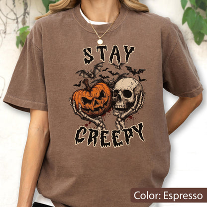 May include: An espresso brown t-shirt featuring a graphic print of a carved orange pumpkin and a human skull held by skeletal hands. Above and below the central illustration, the text reads STAY CREEPY in a black, jagged, distressed font.