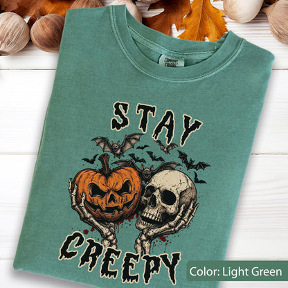 May include: A light green t-shirt featuring a graphic print of two skeletal hands holding a carved jack-o'-lantern and a human skull. Above and below the graphic, the text reads STAY CREEPY in a black, dripping font. The shirt has a crew neckline.