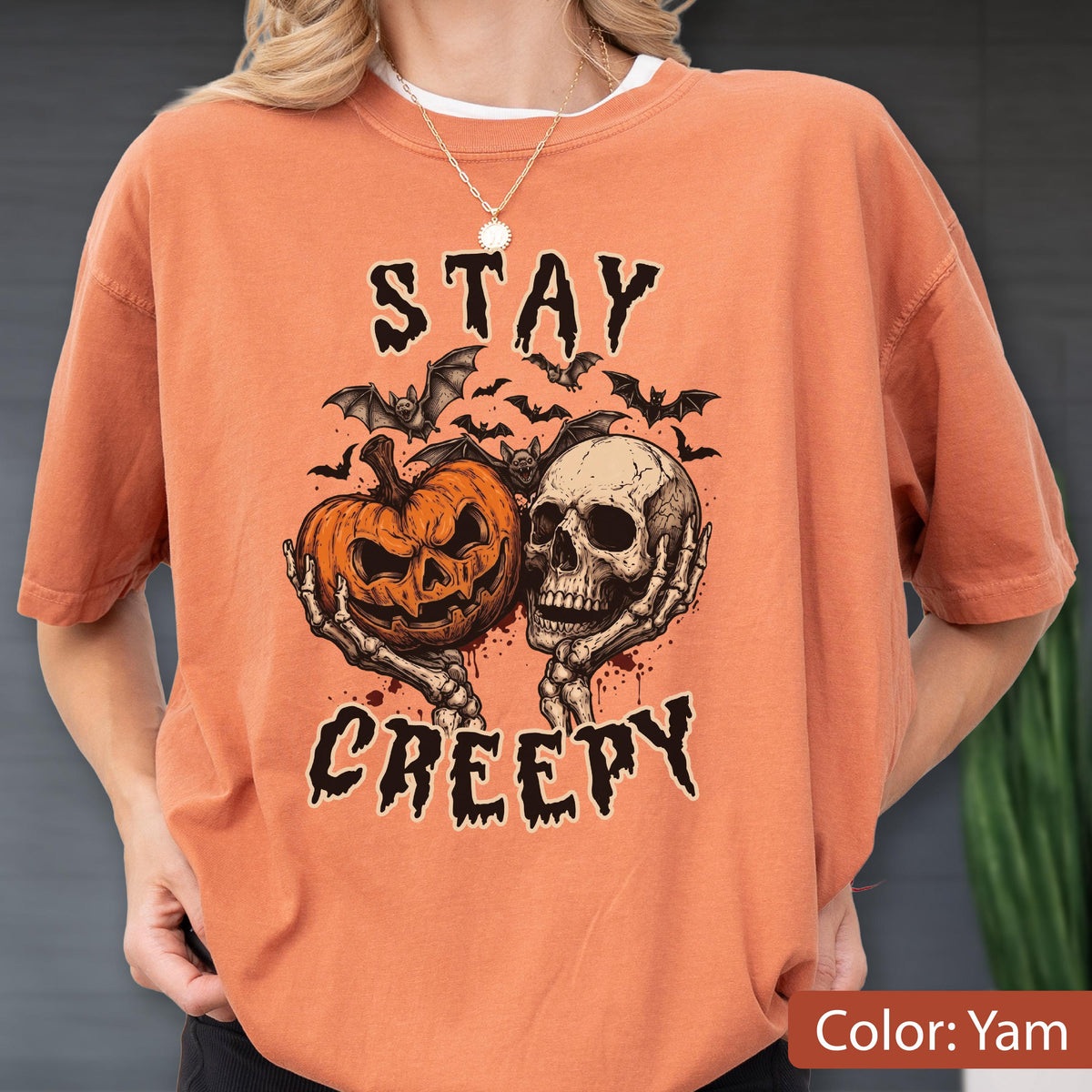 May include: A yam-colored t-shirt featuring a graphic print of a jack-o'-lantern and a human skull held by skeletal hands. Above and below the illustration, the text reads STAY CREEPY in a black, jagged font, surrounded by several small bats.