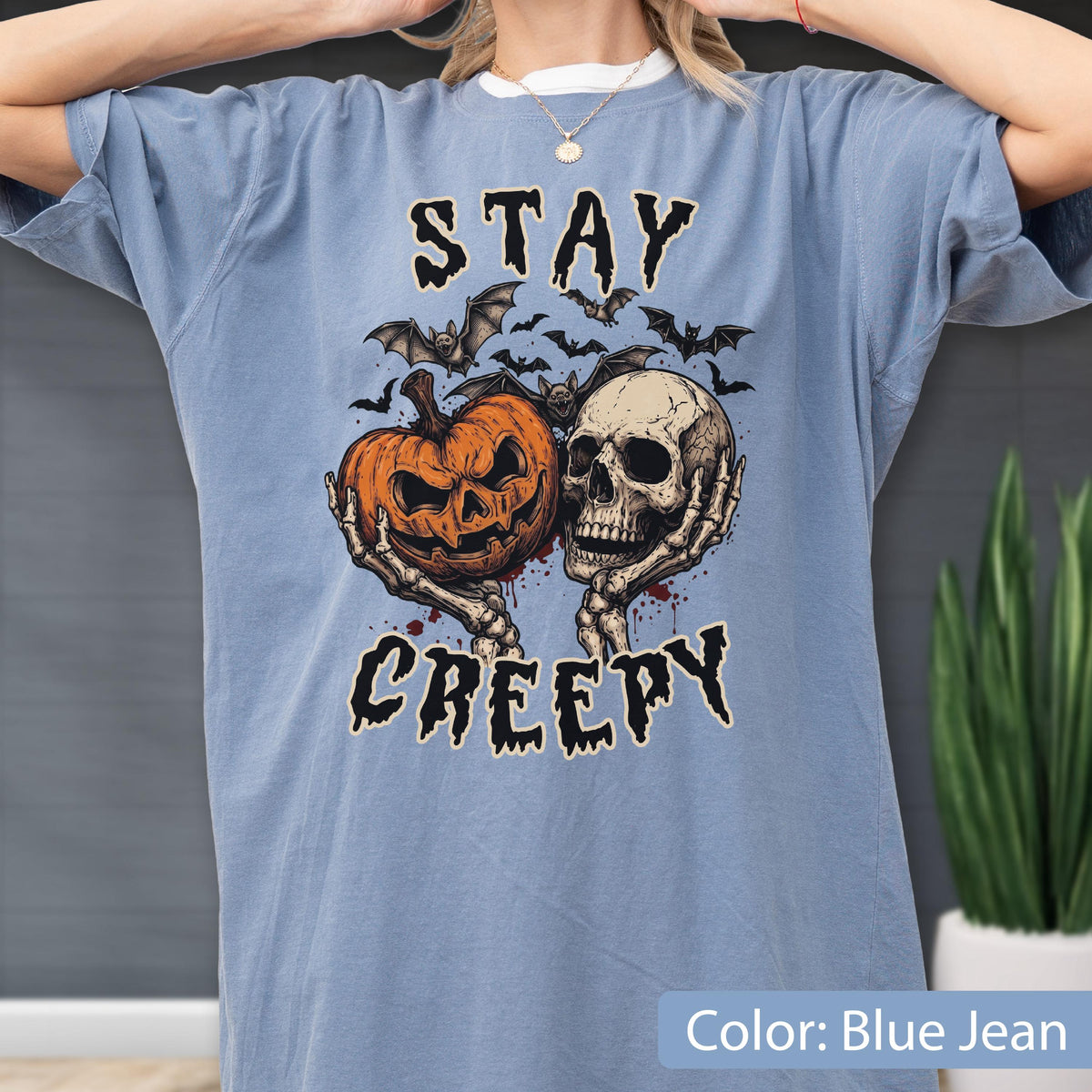 May include: A blue jean colored t-shirt featuring a graphic print of a jack-o'-lantern pumpkin and a human skull held by skeletal hands. Above and below the central illustration, the text reads STAY CREEPY in a jagged, dark font style.