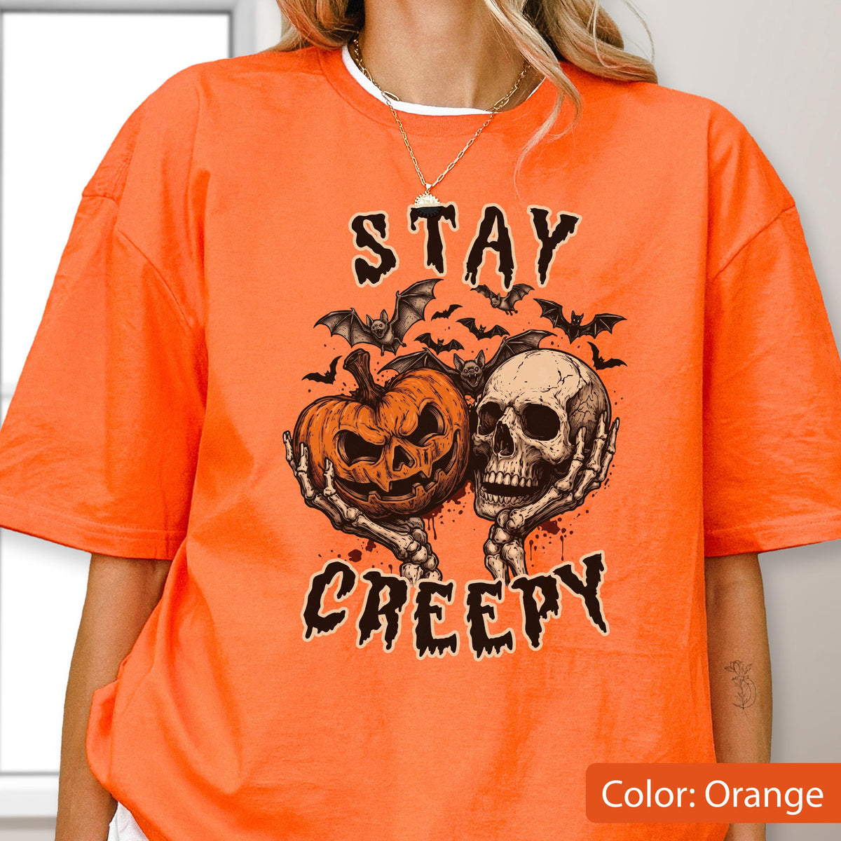 May include: An orange short-sleeve t-shirt featuring a graphic print of a jack-o'-lantern and a human skull held by skeletal hands. Above and below the central illustration, the text reads STAY CREEPY in a black, dripping, jagged font.
