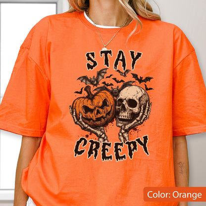 May include: An orange short-sleeve t-shirt featuring a graphic print of a jack-o'-lantern and a human skull held by skeletal hands. Above and below the central illustration, the text reads STAY CREEPY in a black, dripping, jagged font.