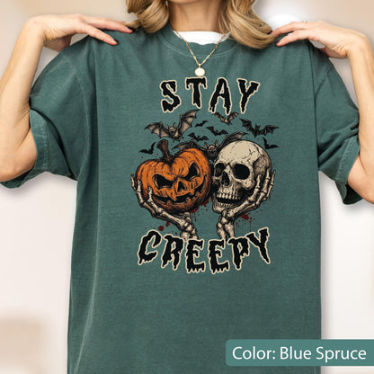 May include: A blue spruce t-shirt featuring a graphic print of a jack-o'-lantern and a human skull held by skeletal hands, surrounded by bats. The text "STAY CREEPY" is printed in a stylized font above and below the central illustration.
