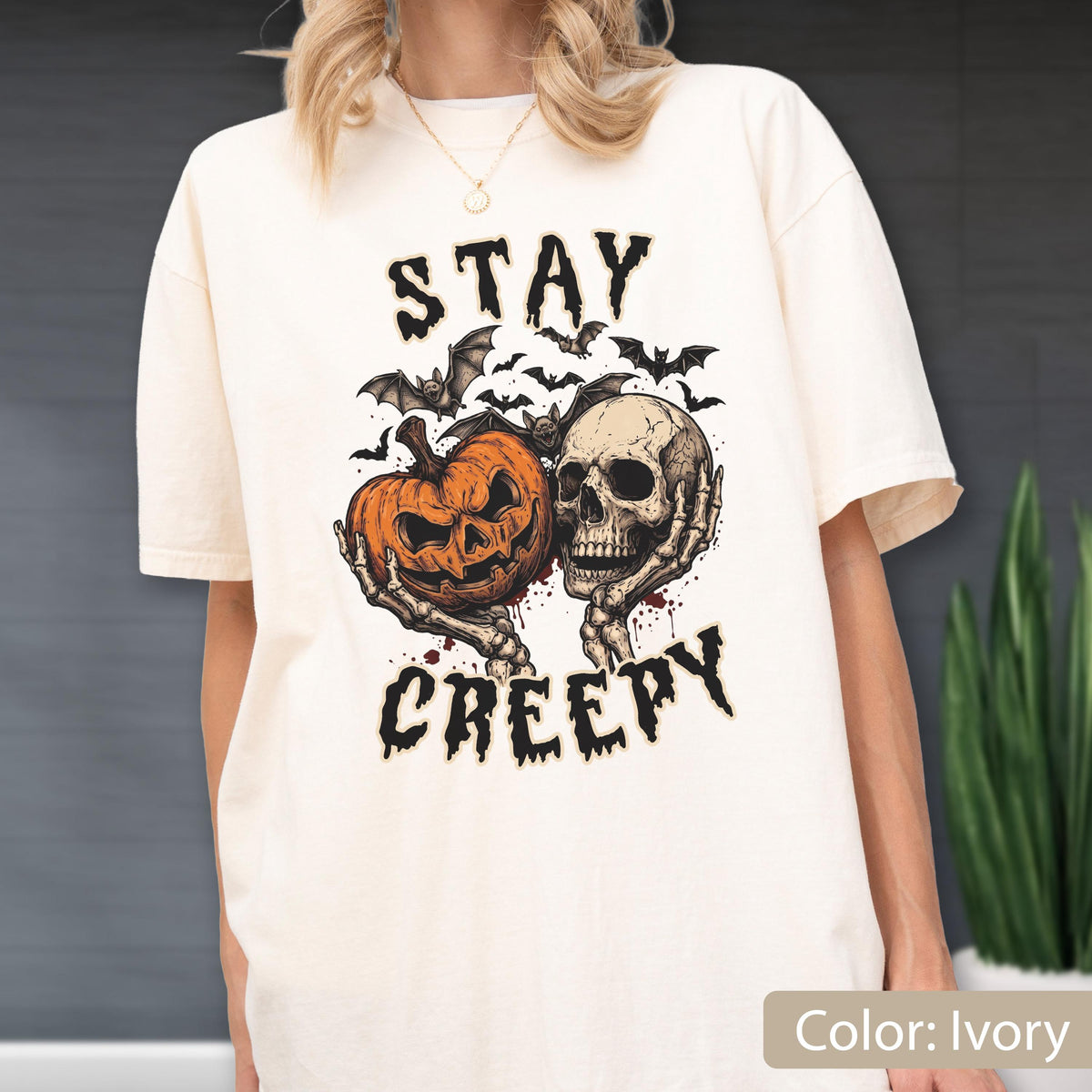 May include: An ivory short-sleeve t-shirt featuring a graphic print of a skeleton holding an orange jack-o'-lantern, surrounded by black bats. The text "STAY CREEPY" is printed in a black, jagged font above and below the central illustration.