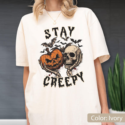 May include: An ivory short-sleeve t-shirt featuring a graphic print of a skeleton holding an orange jack-o'-lantern, surrounded by black bats. The text "STAY CREEPY" is printed in a black, jagged font above and below the central illustration.