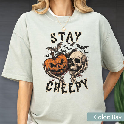 May include: A light bay-colored t-shirt featuring a graphic print of a jack-o'-lantern and a human skull held by skeletal hands. Above and below the illustration, the text reads STAY CREEPY in a black, jagged font, surrounded by small bats.