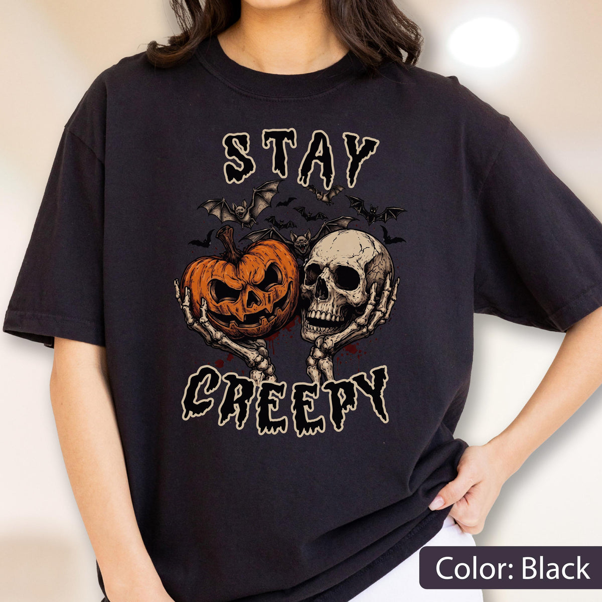 May include: A black t-shirt featuring a graphic print of two skeletal hands holding an orange jack-o'-lantern and a human skull. Above and below the central illustration, the text reads STAY CREEPY in a stylized, dripping font with bats.