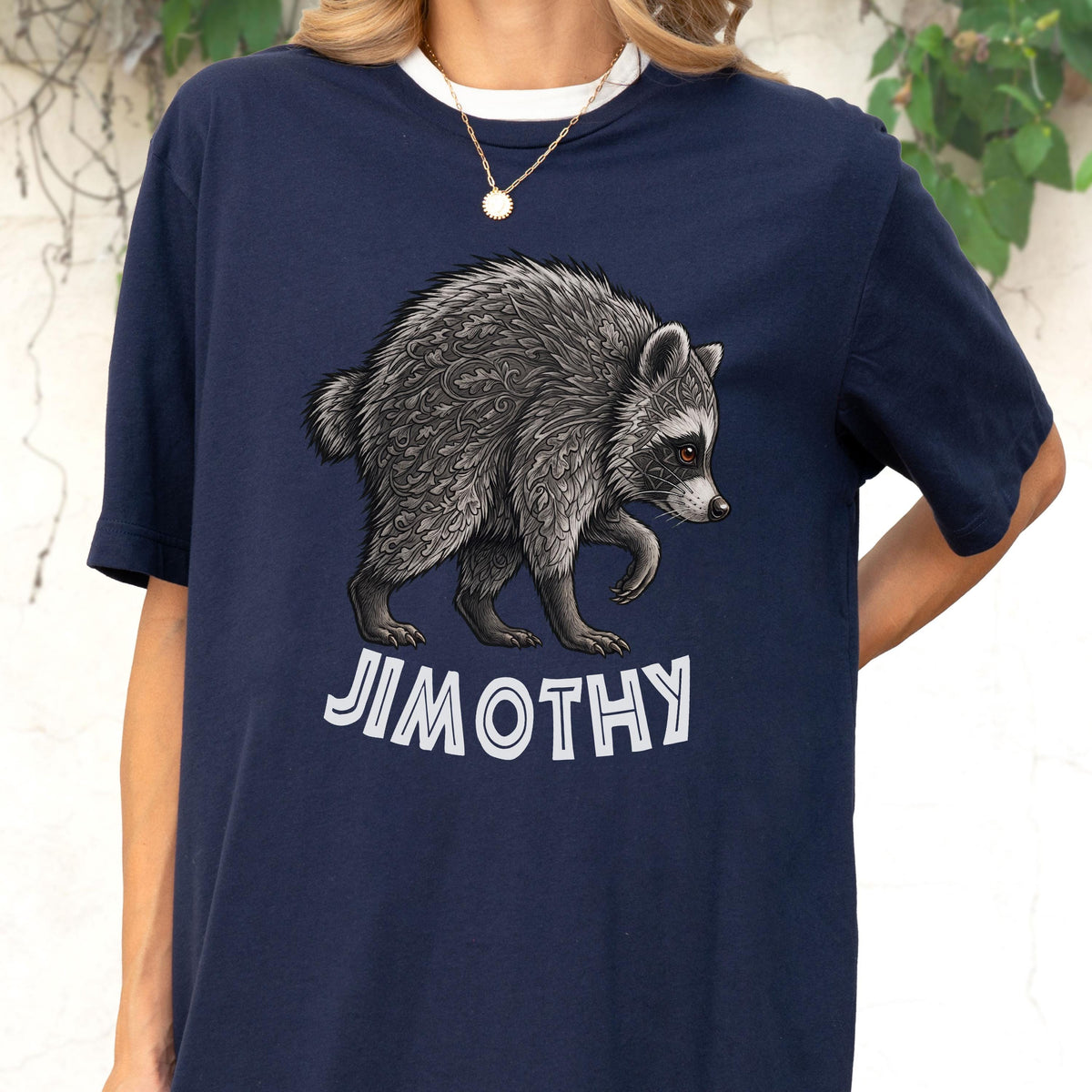 May include: A navy blue t-shirt featuring a graphic illustration of a raccoon with intricate leaf-like patterns in its fur. Below the raccoon, the text JIMOTHY is printed in bold, white, sans-serif capital letters. The shirt has short sleeves.