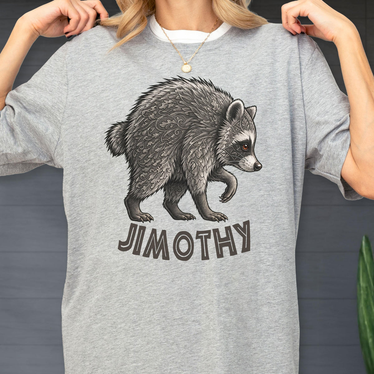 May include: A heather gray t-shirt featuring a detailed illustration of a raccoon with intricate leaf-like patterns in its fur. Below the raccoon, the name JIMOTHY is printed in bold, dark, sans-serif capital letters on the front of the shirt.